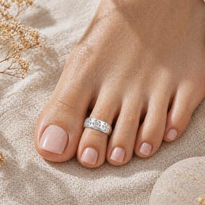 May include: A silver toe ring with a floral design is worn on a foot with a neutral-toned nail polish. The ring is centered on the second toe. The background is a neutral-colored fabric with dried flowers.