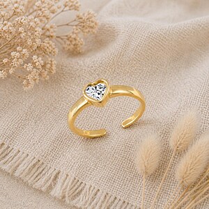Könnte beinhalten: Ein goldfarbener Ring mit einem herzförmigen, klaren Edelstein. Der Ring hat ein offenes Design, das eine leichte Größenanpassung ermöglicht. Der Ring wird auf einem beigefarbenen Stoff mit getrockneten Blumen im Hintergrund präsentiert.