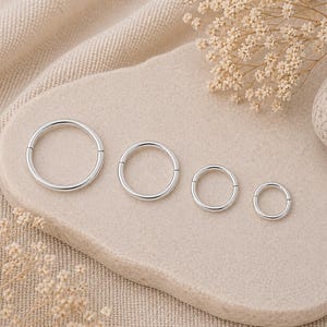 May include: Four silver-colored, hinged hoop earrings of varying sizes are arranged on a light beige, organic-shaped stone. The earrings are circular, with a simple, polished design. The background is a textured, light beige fabric.