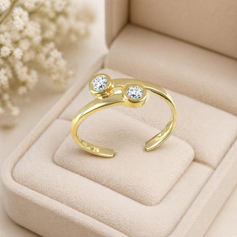 Pu&ograve; includere: Un anello dorato con due gemme rotonde e trasparenti. L'anello ha un design aperto unico ed &egrave; presentato in un portagioie beige. Le gemme sono incastonate in uno stile moderno e minimalista.