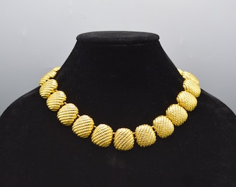 May include: A gold-toned necklace with a series of textured, round, linked elements. The necklace is displayed on a black mannequin. The design features a repeating pattern of ribbed, spherical shapes.