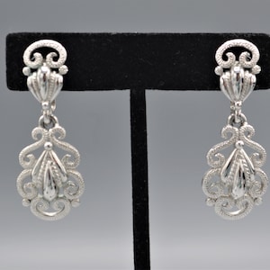 May include: A pair of silver dangle earrings with a filigree design. The earrings have a teardrop shape and are accented with a small, round bead at the top.