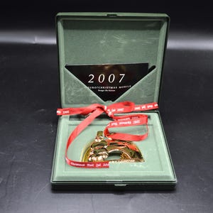 May include: A gold-colored Christmas ornament in a green box, with a red ribbon. The ornament is in the shape of a bridge. The box contains a card with the year "2007" printed on it. The ribbon has the year "2007" printed on it.