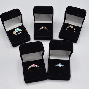 May include: Five silver-toned rings, each featuring a different colored gemstone, are displayed in individual black velvet jewelry boxes. The rings include a square blue stone, a round red stone, a small green stone, a pink stone, and a blue heart-shaped stone.