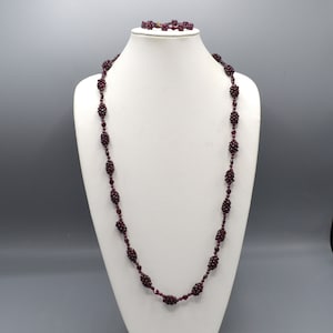 May include: A long necklace and matching bracelet set featuring deep dark red, faceted, clustered bead segments separated by smaller individual beads. The jewelry pieces are displayed on a white neck form, showcasing the uniform, repeating pattern.