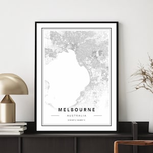 Pode incluir: Uma impressão de mapa preto e branco de Melbourne, Austrália, em uma moldura preta, pendurada em uma parede branca acima de uma mesa de console marrom escura com uma lâmpada dourada e uma cadeira tecida.