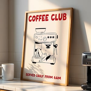 May include: A framed print with the words "COFFEE CLUB" in red, featuring a black and white illustration of an espresso machine with a person relaxing on it. A white mug and a black coffee maker are also visible.