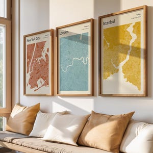 May include: Three framed city map prints of New York City, London, and Istanbul hang above a bench with pillows. The New York City map is red, London is blue, and Istanbul is yellow. The bench has beige cushions and several pillows.