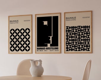 May include: Three framed black and white Bauhaus art prints with geometric designs and text that reads "Bauhaus-Ausstellung Weimar Juli - Sept 1923". The prints are hanging on a white wall above a wooden table with a white ceramic pitcher and four beige chairs.