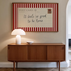 May include: A framed postcard print with red and white stripes and the text "It feels so good to be home" hangs above a wooden cabinet. A white lamp and a small white bowl sit on the cabinet.