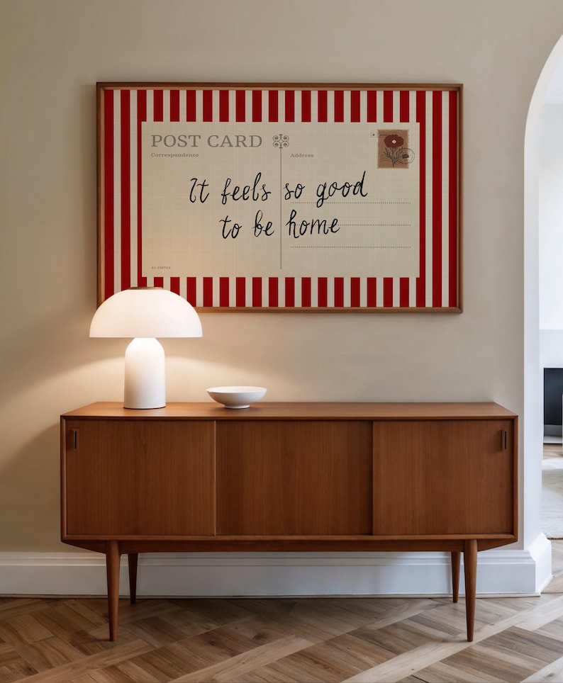 May include: A framed postcard print with red and white stripes and the text "It feels so good to be home" hangs above a wooden cabinet. A white lamp and a small white bowl sit on the cabinet.