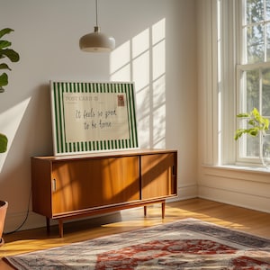 May include: A framed postcard print with the text "It feels so good to be home" is displayed on a wooden cabinet. The postcard has a green and white striped border and a postage stamp design. The cabinet is in a room with a window and a patterned rug.
