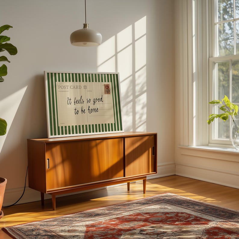 May include: A framed postcard print with the text "It feels so good to be home" is displayed on a wooden cabinet. The postcard has a green and white striped border and a postage stamp design. The cabinet is in a room with a window and a patterned rug.