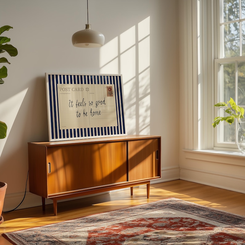May include: A framed postcard print with a navy blue and white striped border. The postcard reads "It feels so good to be home" in a handwritten font. The print is displayed on a wooden cabinet in a well-lit room.