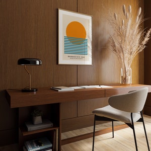 May include: A minimalist home office with a wooden desk, a black lamp, a beige chair, and a framed print with a geometric design of a sun and blue stripes. The print has the text "Bauhaus 1919-1933, School of Art and Crafts". A vase with dried pampas grass is on the desk.