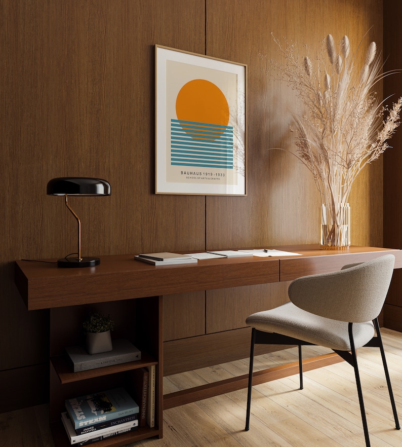 May include: A minimalist home office with a wooden desk, a black lamp, a beige chair, and a framed print with a geometric design of a sun and blue stripes. The print has the text "Bauhaus 1919-1933, School of Art and Crafts". A vase with dried pampas grass is on the desk.