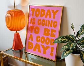 May include: A framed pink poster with the text "TODAY IS GOING TO BE A GOOD DAY!" in orange letters. A gold-framed poster is propped against a wall next to an orange lamp and a potted plant.