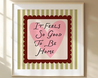 Könnte beinhalten: Gerahmter Kunstdruck mit einem rosa Herzen und dem Schriftzug "It Feels So Good To Be Home" in schwarzer Schrift. Das Herz befindet sich auf einem cremefarbenen Hintergrund, umrahmt von einem dunkelroten, gewellten Rand, auf einem grün-cremefarbenen Streifenhintergrund.