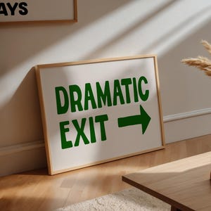 May include: A framed white sign with the words "DRAMATIC EXIT" in green, and a green arrow pointing right. The sign is on a wooden floor. Another framed sign is visible in the background.