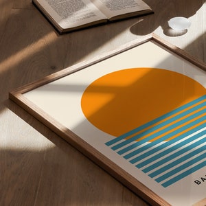 May include: A framed print with a minimalist design featuring a large orange circle representing the sun, partially obscured by a series of teal blue horizontal stripes, suggesting the ocean. The text "BA" is visible at the bottom of the print.