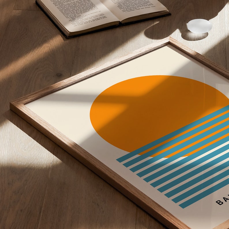 May include: A framed print with a minimalist design featuring a large orange circle representing the sun, partially obscured by a series of teal blue horizontal stripes, suggesting the ocean. The text "BA" is visible at the bottom of the print.
