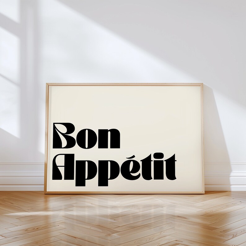 May include: A framed art print with the words "Bon App&eacute;tit" in bold, black, stylized lettering. The background is a soft cream color, and the frame is a light wood tone. The print is displayed against a white wall and a wooden floor.