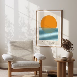May include: A framed print with a minimalist geometric design featuring a large orange circle over a series of blue and teal horizontal lines. The text "BAUHAUS 1919 - 1933 SCHOOL OF ARTS & CRAFTS" is printed below the design.