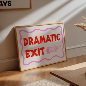 Puede incluir: Un cuadro enmarcado con las palabras "DRAMATIC EXIT" en rojo, con un borde ondulado rosa y una ilustración de una mano señalando. La impresión está sobre un fondo blanco y en un marco de madera clara.