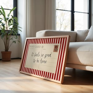 May include: A framed postcard print with a red and white striped border. The postcard has the text "It feels so good to be home" in black script. A small floral stamp is in the upper right corner. The frame is made of light-colored wood.