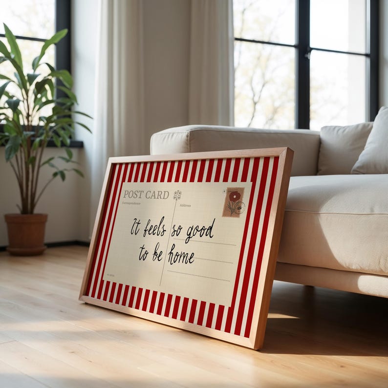 May include: A framed postcard print with a red and white striped border. The postcard has the text "It feels so good to be home" in black script. A small floral stamp is in the upper right corner. The frame is made of light-colored wood.