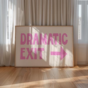 May include: A framed art print with the words "DRAMATIC EXIT" in pink, along with a pink arrow pointing right. The artwork is set against a cream background and framed in a light wood. The print is displayed on a wooden floor.