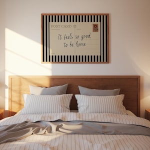 May include: A framed wall art piece with a postcard design. The postcard reads "It feels so good to be home" in script font. The frame has a black and white striped border. The art hangs above a bed with striped pillows and a gray blanket.