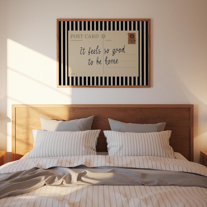 May include: A framed wall art piece with a postcard design. The postcard reads "It feels so good to be home" in script font. The frame has a black and white striped border. The art hangs above a bed with striped pillows and a gray blanket.