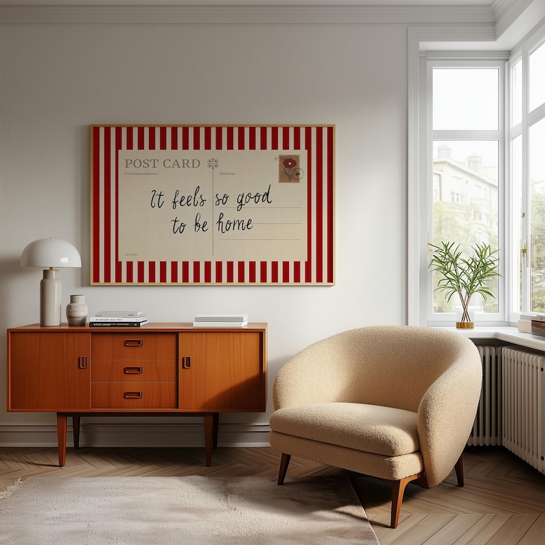 May include: A framed postcard print with red and white stripes and the text "It feels so good to be home." The print hangs above a mid-century modern wooden cabinet and a beige armchair.