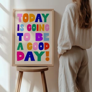 May include: A framed art print with the words "TODAY IS GOING TO BE A GOOD DAY!" in colorful block letters. The print is in a light wood frame and is displayed on a small wooden stool. The background is a neutral color.