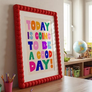 May include: A framed print with the words "TODAY IS GOING TO BE A GOOD DAY!" in colorful block letters. The frame is red with a textured, rounded design. The print is displayed on a wooden surface.