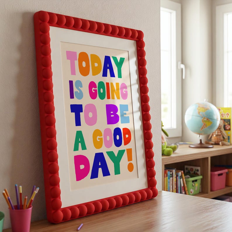 May include: A framed print with the words "TODAY IS GOING TO BE A GOOD DAY!" in colorful block letters. The frame is red with a textured, rounded design. The print is displayed on a wooden surface.