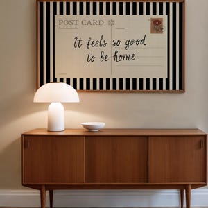 May include: A mid-century modern wooden sideboard with a white lamp and a small white bowl on top. A framed postcard with black and white stripes and the text "It feels so good to be home" hangs on the wall.