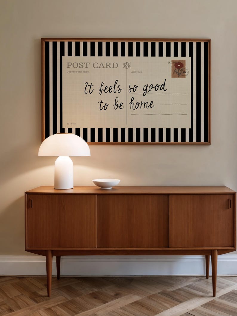May include: A mid-century modern wooden sideboard with a white lamp and a small white bowl on top. A framed postcard with black and white stripes and the text "It feels so good to be home" hangs on the wall.