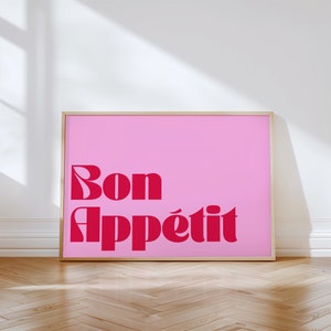 May include: A framed pink art print with the words "Bon App&eacute;tit" in a bold, red, retro font. The artwork is displayed on a light-colored wall, with a wooden floor in the foreground.