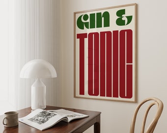 Puede incluir: Un póster enmarcado con las palabras "Gin & Tonic" en letras de bloque verdes y rojas. El póster está en un marco de madera clara y cuelga de una pared de color crema. Una lámpara blanca, una taza y una revista abierta están sobre un escritorio de madera oscura.