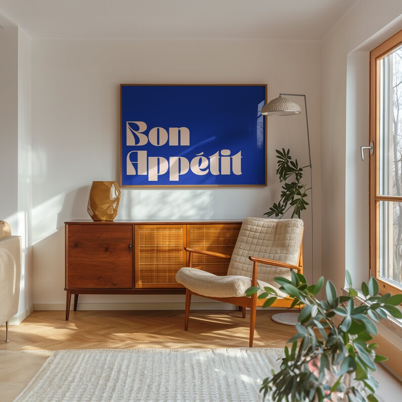 May include: A room interior featuring a blue wall art with the text "Bon App&eacute;tit" in a light beige font. The room includes a wooden cabinet, a beige armchair, a floor lamp, and a decorative vase. The floor is made of wood.