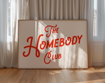 May include: A framed art print with the words "The Homebody Club" in red script on a cream background. The artwork is displayed on a wooden floor, with white curtains and a window in the background.