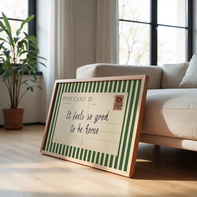 May include: A framed postcard with a wooden frame, featuring green and white stripes and the text "It feels so good to be home." The postcard has a vintage aesthetic with a floral stamp.