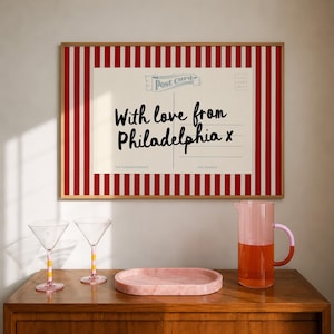 Op de afbeelding: Een ingelijste kunstprint met een ansichtkaartontwerp met de tekst 'With love from Philadelphia x' tegen een rood-witte verticaal gestreepte achtergrond, vergezeld door twee martiniglazen, een roze ovaal dienblad en een tweekleurige karaf.