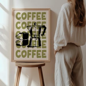 May include: A framed print with a coffee theme. The artwork features the word "COFFEE" in olive green block letters, along with a black and white illustration of a coffee maker and two stacked coffee cups. The frame is a light wood color.