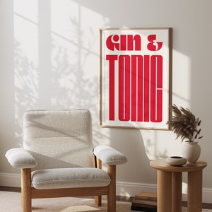May include: A framed print with the text "Gin & Tonic" in bold, red letters on a white background. The print is hanging on a white wall in a room with a light wood chair and a small table.