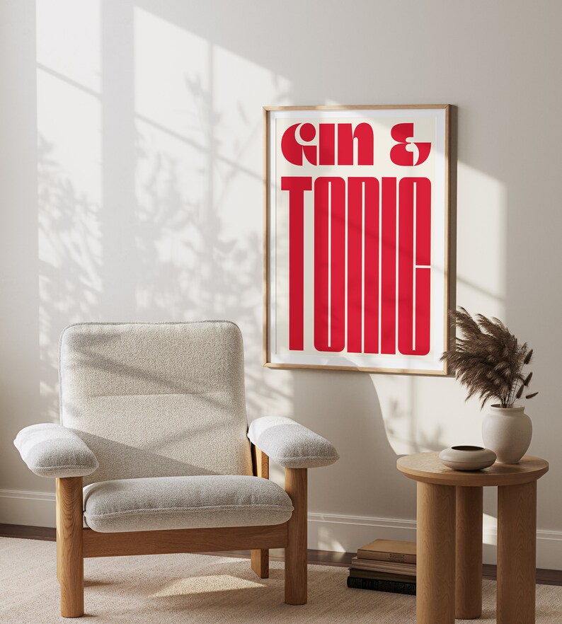 May include: A framed print with the text "Gin & Tonic" in bold, red letters on a white background. The print is hanging on a white wall in a room with a light wood chair and a small table.