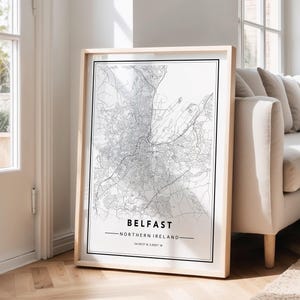 May include: A framed monochrome map of Belfast, Northern Ireland, showing the city's layout, with the name and coordinates. The map is in a light wood frame, positioned against a wall near a window and a beige sofa.