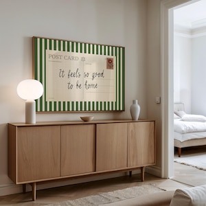 May include: A wooden sideboard with four doors and a light-colored finish. A framed postcard with green and white stripes and the text "It feels so good to be home" hangs above. A white lamp and vase are on the sideboard.